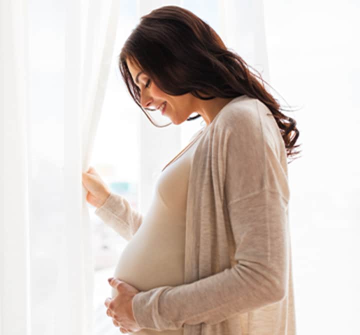 Mulher grávida feliz olhando para a barriga em pé ao lado da janela 